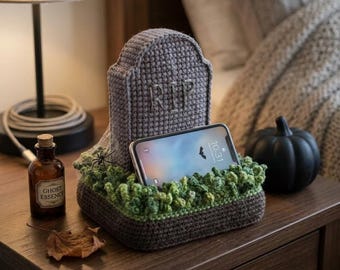 May include: A gray crocheted tombstone phone stand featuring the letters RIP stitched on the front. The base is decorated with textured green yarn to resemble grass and includes a small black spider accent. A smartphone rests in the stand.