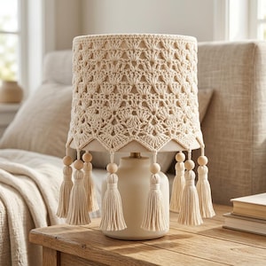 May include: A table lamp featuring a cream-colored ceramic base and a cylindrical lampshade covered in an intricate, off-white crocheted lace overlay. The shade is finished with a scalloped bottom edge adorned with hanging wooden beads and tassels.