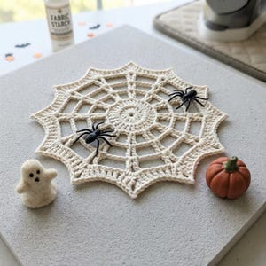 May include: A cream-coloured crocheted spider web doily featuring two black plastic spiders, a small white felted ghost figure, and a miniature orange pumpkin. The circular web design has a central point with radiating spokes and concentric rings.
