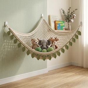 May include: A cream-colored crocheted toy hammock featuring a diamond-patterned mesh and decorative green leaf accents along the bottom edge. The hammock is suspended by three white wall hooks and contains a plush bear, elephant, and deer.
