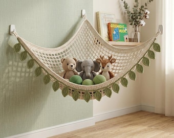 May include: A cream-colored crocheted toy hammock featuring a diamond-patterned mesh and decorative green leaf accents along the bottom edge. The hammock is suspended by three white wall hooks and contains a plush bear, elephant, and deer.