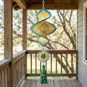 Puede incluir: Un adorno de ganchillo en espiral colgante con capas concéntricas de hilo amarillo, azul claro y verde. Debajo de la espiral cuelga un motivo floral de seis pétalos en colores a juego, decorado con pequeñas cuentas y una borla de hilo verde.