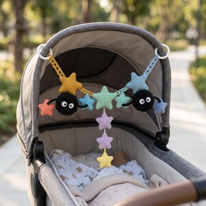 May include: A crochet stroller toy featuring two black fuzzy creatures with large white eyes, connected by a string of multi-colored stars in yellow, light blue, mint green, and lavender, suspended from two white plastic rings.