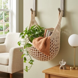 May include: A cream-colored, hand-crocheted hanging storage basket with a diamond-patterned mesh design. The basket is suspended from two wooden wall hooks and contains a green leafy plant, a folded orange textile, and three small books.