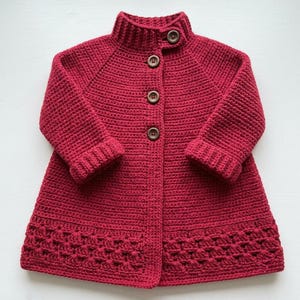May include: A deep red crochet cardigan featuring a high collar, long sleeves with ribbed cuffs, and a button-down front with four dark brown buttons. The garment has a textured stitch pattern throughout and a decorative openwork border at the hem.