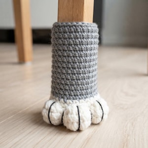 May include: A gray crocheted furniture leg cover shaped like an animal paw. The cylindrical upper section is made of gray yarn, while the base features four white crocheted toe pads, each accented with a thin black vertical line for detail.