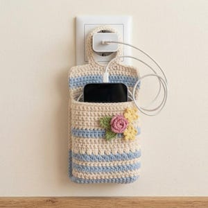 Könnte beinhalten: Ein beigefarbener und hellblau gestreifter Handyhalter aus Häkelarbeit, der an einem weißen Wandladegerät hängt. Der Halter ist mit einer kleinen gehäkelten rosa Rose mit grünen Blättern und zwei gelben Blumen verziert und enthält ein schwarzes Smartphone.