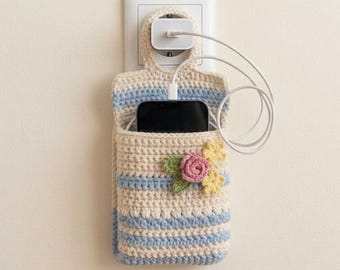 May include: A beige and light blue striped crochet phone holder hanging from a white wall charger. The holder features a small pink crocheted rose with green leaves and two yellow flowers on the front, containing a black smartphone.
