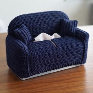 May include: A navy blue crochet tissue box cover shaped like a miniature sofa. The cover features a high backrest, two side armrests, and two small square cushions on each side. White facial tissues are visible protruding from the center opening.