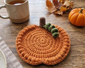 May include: A round, orange crochet coaster shaped like a pumpkin. The coaster features a textured, spiraled stitch pattern, a small brown crocheted stem at the top, and a coiled green crocheted vine accent attached next to the stem.
