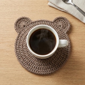 May include: A brown crocheted coaster shaped like a bear head with rounded ears. A white ceramic mug filled with dark coffee sits in the center of the coaster. The coaster features a dense, circular stitch pattern made from brown yarn.