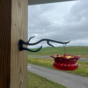 May include: A black metal wall-mounted bracket shaped like a tree branch, attached to a wooden post. A red plastic bird feeder with a scalloped top edge and a clear ribbed base is suspended from the end of the bracket by a silver metal hook.