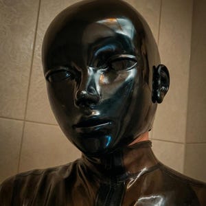 May include: A glossy black full-head hood with a smooth, reflective surface covering the entire head and neck. The hood features molded facial contours, including defined eyes, a nose, and a mouth area, with a vertical zipper running down the front.