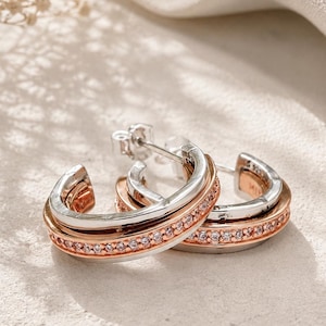 Puede incluir: Un par de pendientes de aro bicolor con una banda exterior plateada y una banda interior de color oro rosa incrustada con una hilera de pequeñas piedras transparentes de corte redondo. Los pendientes tienen cierre de perno y sello 925.