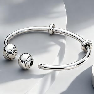Puede incluir: Una pulsera rígida abierta de plata pulida con una banda circular lisa. La pulsera incluye dos extremos esféricos grabados con el texto PANDORA, junto con dos cuentas espaciadoras deslizantes adicionales situadas a lo largo del marco.