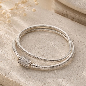 Puede incluir: Una pulsera de cadena de serpiente en tono plateado con un diseño de doble vuelta y un cierre cilíndrico incrustado con pequeñas piedras claras y brillantes. La pulsera tiene una apariencia tejida texturizada y un acabado metálico pulido.