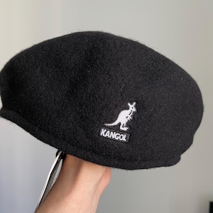 May include: A black wool-blend flat cap featuring a rounded crown and a short, stiff brim. The side of the cap displays an embroidered white kangaroo logo above the text KANGOL in a black rectangular patch with white border stitching.