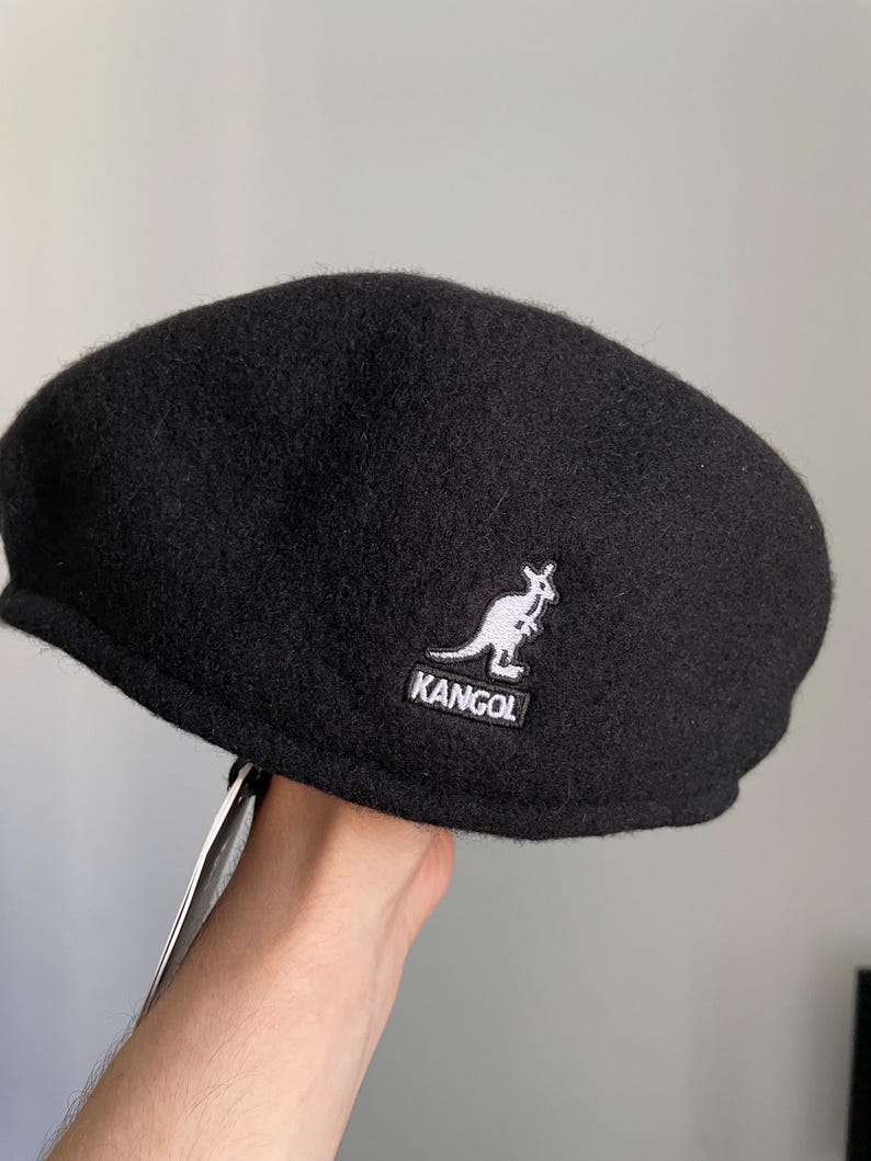 May include: A black wool-blend flat cap featuring a rounded crown and a short, stiff brim. The side of the cap displays an embroidered white kangaroo logo above the text KANGOL in a black rectangular patch with white border stitching.