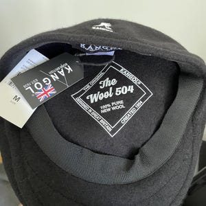 May include: A black Kangol 504 cap made of 100% pure new wool. The interior features a white label with the text The Wool 504, Designed in Great Britain, Created 1954. A black hang tag with the Kangol logo and a Union Jack flag is attached.