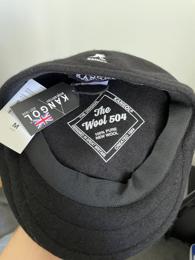 May include: A black Kangol 504 cap made of 100% pure new wool. The interior features a white label with the text The Wool 504, Designed in Great Britain, Created 1954. A black hang tag with the Kangol logo and a Union Jack flag is attached.