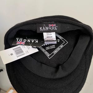 May include: A black Kangol wool cap with a black inner lining and a black sweatband. The interior features a black label with the Kangol logo, a Union Jack, and the text Born British Est. 1938. A white tag indicates 100% pure new wool and size medium.