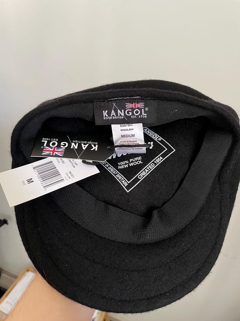 May include: A black Kangol wool cap with a black inner lining and a black sweatband. The interior features a black label with the Kangol logo, a Union Jack, and the text Born British Est. 1938. A white tag indicates 100% pure new wool and size medium.