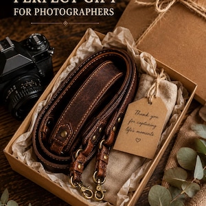 May include: A dark brown leather camera strap with brass hardware coiled inside a cardboard gift box. The box contains a beige fabric pouch with a brown paper tag that reads Thank you for capturing life's moments with a heart symbol.