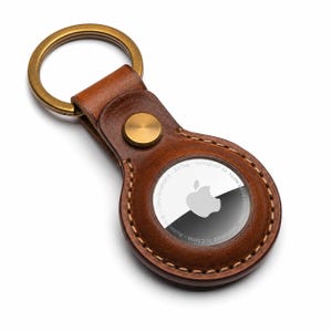 May include: A brown leather keychain holder featuring a circular cutout for an Apple AirTag. The holder is secured with a brass-colored metal snap button and includes a matching brass-colored metal split ring for keys, with visible stitching.