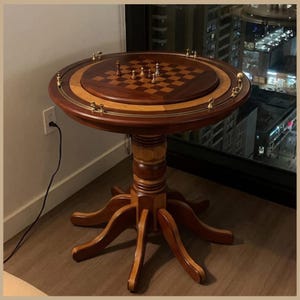 May include: A round wooden game table featuring a built-in chessboard on the top surface. The table has a dark brown finish with lighter wood accents on the pedestal base and legs. A brass rail with small decorative metal spheres encircles the top.