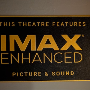 May include: A rectangular black sign with gold lettering that reads THIS THEATRE FEATURES IMAX ENHANCED PICTURE & SOUND. The text is arranged in three lines with the word IMAX in a large, bold font, centered on a dark, textured background.