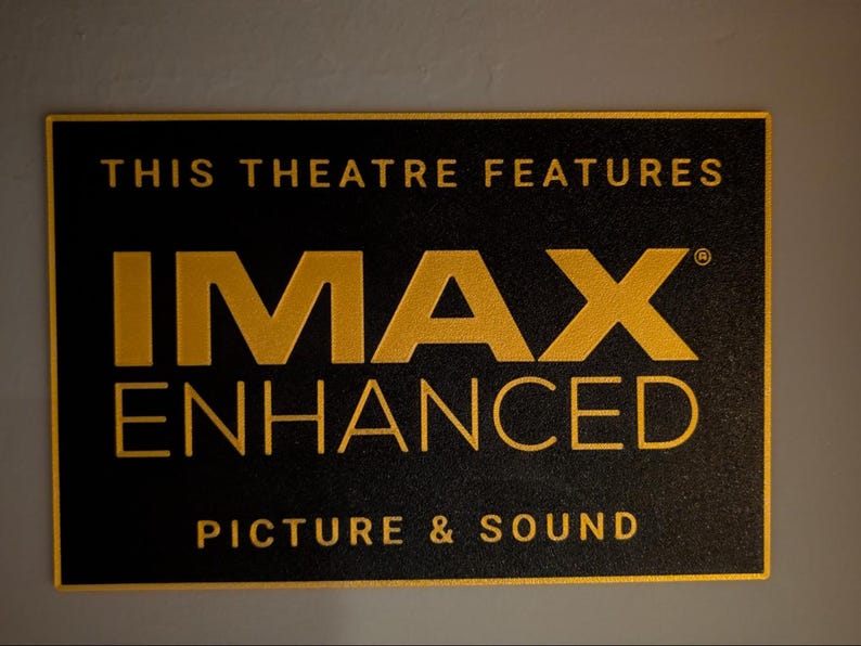 May include: A rectangular black sign with gold lettering that reads THIS THEATRE FEATURES IMAX ENHANCED PICTURE & SOUND. The text is arranged in three lines with the word IMAX in a large, bold font, centered on a dark, textured background.