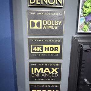May include: A vertical arrangement of five rectangular black plaques with gold lettering. The plaques display the text: THIS THEATRE IS POWERED BY DENON, THIS THEATRE FEATURES DOLBY ATMOS, THIS THEATRE FEATURES 4K HDR HIGH DYNAMIC RANGE, THIS THEATRE FEATURES IMAX ENHANCED PICTURE & SOUND, and THIS THEATRE FEATURES EPSON PROJECTION SYSTEM.