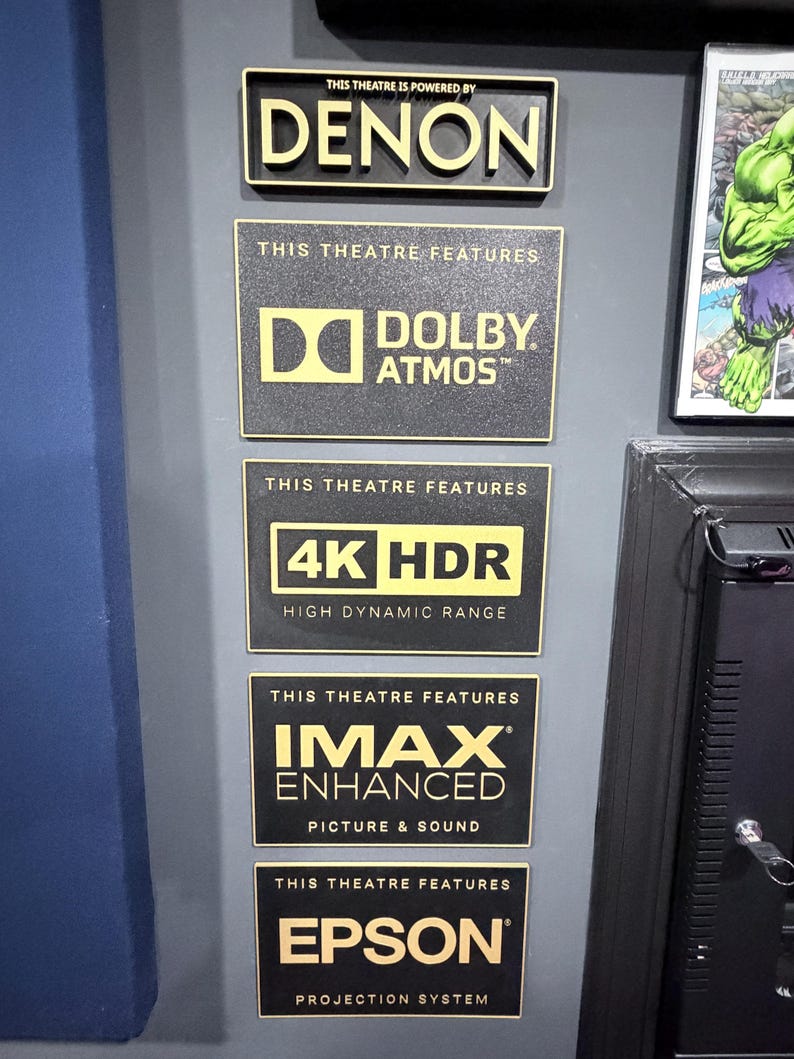 May include: A vertical arrangement of five rectangular black plaques with gold lettering. The plaques display the text: THIS THEATRE IS POWERED BY DENON, THIS THEATRE FEATURES DOLBY ATMOS, THIS THEATRE FEATURES 4K HDR HIGH DYNAMIC RANGE, THIS THEATRE FEATURES IMAX ENHANCED PICTURE & SOUND, and THIS THEATRE FEATURES EPSON PROJECTION SYSTEM.