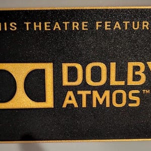 May include: A rectangular black sign with a textured surface and a gold border. The text reads THIS THEATRE FEATURES in gold, followed by the gold Dolby Atmos logo and the word ATMOS in gold lettering. The sign has a matte finish throughout.