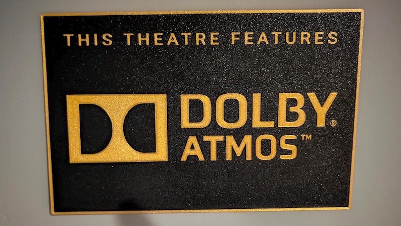 May include: A rectangular black sign with a textured surface and a gold border. The text reads THIS THEATRE FEATURES in gold, followed by the gold Dolby Atmos logo and the word ATMOS in gold lettering. The sign has a matte finish throughout.