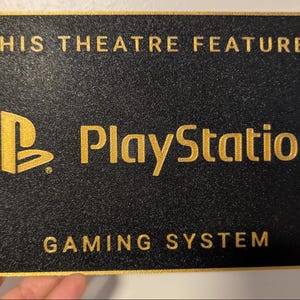 May include: A rectangular black sign with a textured surface and gold border. The text reads THIS THEATRE FEATURES in gold, followed by the PlayStation logo and the word PlayStation, and the text GAMING SYSTEM at the bottom in gold lettering.