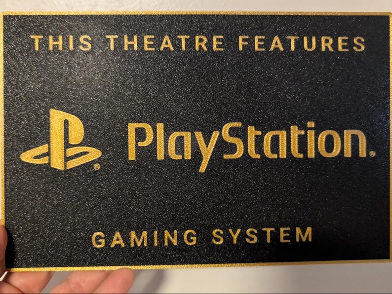 May include: A rectangular black sign with a textured surface and gold border. The text reads THIS THEATRE FEATURES in gold, followed by the PlayStation logo and the word PlayStation, and the text GAMING SYSTEM at the bottom in gold lettering.