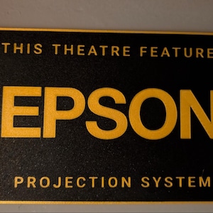 May include: A rectangular black sign with a gold border and gold lettering. The text reads THIS THEATRE FEATURES in the top section, EPSON in the center, and PROJECTION SYSTEM in the bottom section. The surface has a textured, matte finish.