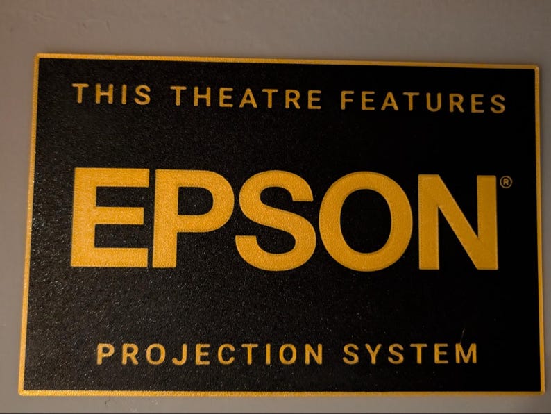 May include: A rectangular black sign with a gold border and gold lettering. The text reads THIS THEATRE FEATURES in the top section, EPSON in the center, and PROJECTION SYSTEM in the bottom section. The surface has a textured, matte finish.