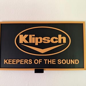 May include: A rectangular wall sign featuring a black background with a light orange frame. The Klipsch logo is centered in light orange, with the text KEEPERS OF THE SOUND displayed in light orange lettering underneath the logo on the black surface.
