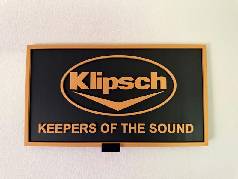 May include: A rectangular wall sign featuring a black background with a light orange frame. The Klipsch logo is centered in light orange, with the text KEEPERS OF THE SOUND displayed in light orange lettering underneath the logo on the black surface.