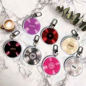 Puede incluir: Un conjunto de siete llaveros circulares diseñados como discos de vinilo, cada uno con un color diferente y una etiqueta central con los nombres Jung Kook, V, j-hope, JIMIN, RM, JIN y SUGA. Cada llavero tiene un cierre de metal plateado.