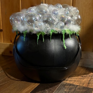 May include: A black plastic cauldron featuring a handle and a decorative top layer of white fiber fill, iridescent clear spheres, and bright green liquid-like drips cascading over the rim of the container, creating a bubbling potion effect.