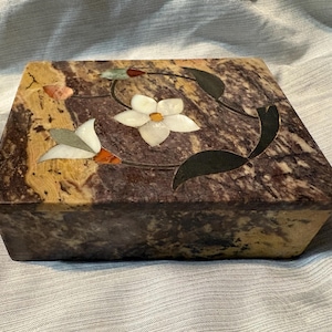 May include: A rectangular stone box featuring a polished dark brown and tan marbled surface. The lid is inlaid with a floral design composed of white, green, and orange stone pieces, depicting a central flower with surrounding leaves and buds.