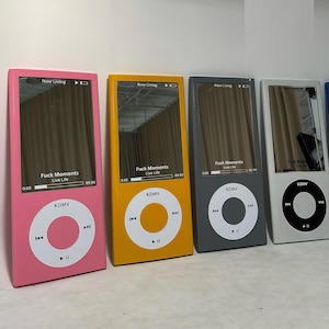 May include: Four rectangular wall mirrors shaped like vintage digital music players in pink, yellow, gray, and white. Each mirror features a screen display with the text Now Living, Fuck Moments, and Live Life, plus a circular control interface.