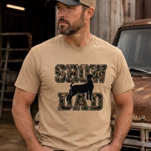Può includere: Una t-shirt a maniche corte e girocollo color beige con la scritta SHOW DAD in un carattere a blocchi con riempimento a motivo mimetico. Una sagoma nera di una capra è posizionata tra le parole SHOW e DAD sulla parte anteriore.