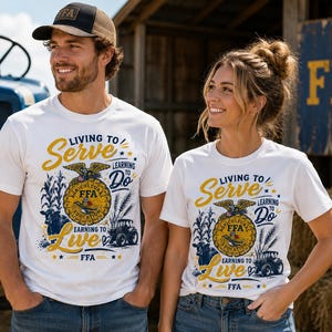 May include: White short-sleeve t-shirts featuring a graphic print with the text Living to Serve, Learning to Do, Earning to Live, and FFA. The design includes a central emblem with an eagle, a tractor, a cow, and stalks of corn in yellow and blue.
