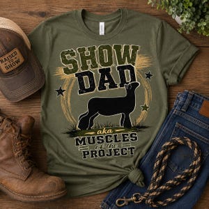 Può includere: Una t-shirt verde oliva con la sagoma nera di un agnello al centro. Il testo recita SHOW DAD in grandi lettere sopra l'agnello e aka MUSCLES of the PROJECT sotto. Il design include piccole stelle e una pennellata circolare.
