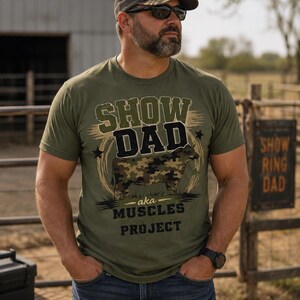 Può includere: Una t-shirt a maniche corte verde oliva con la grafica di una mucca con motivo mimetico. Il testo recita SHOW DAD, aka MUSCLES PROJECT. Il design include un elemento circolare sullo sfondo e due piccole stelle ai lati della grafica.