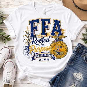 May include: A white short-sleeve t-shirt featuring a graphic with the text FFA, Rooted in Purpose, Growing Leaders, and Est. 1929. The design includes a corn stalk, an eagle emblem, and blue and gold lettering on a white fabric background.